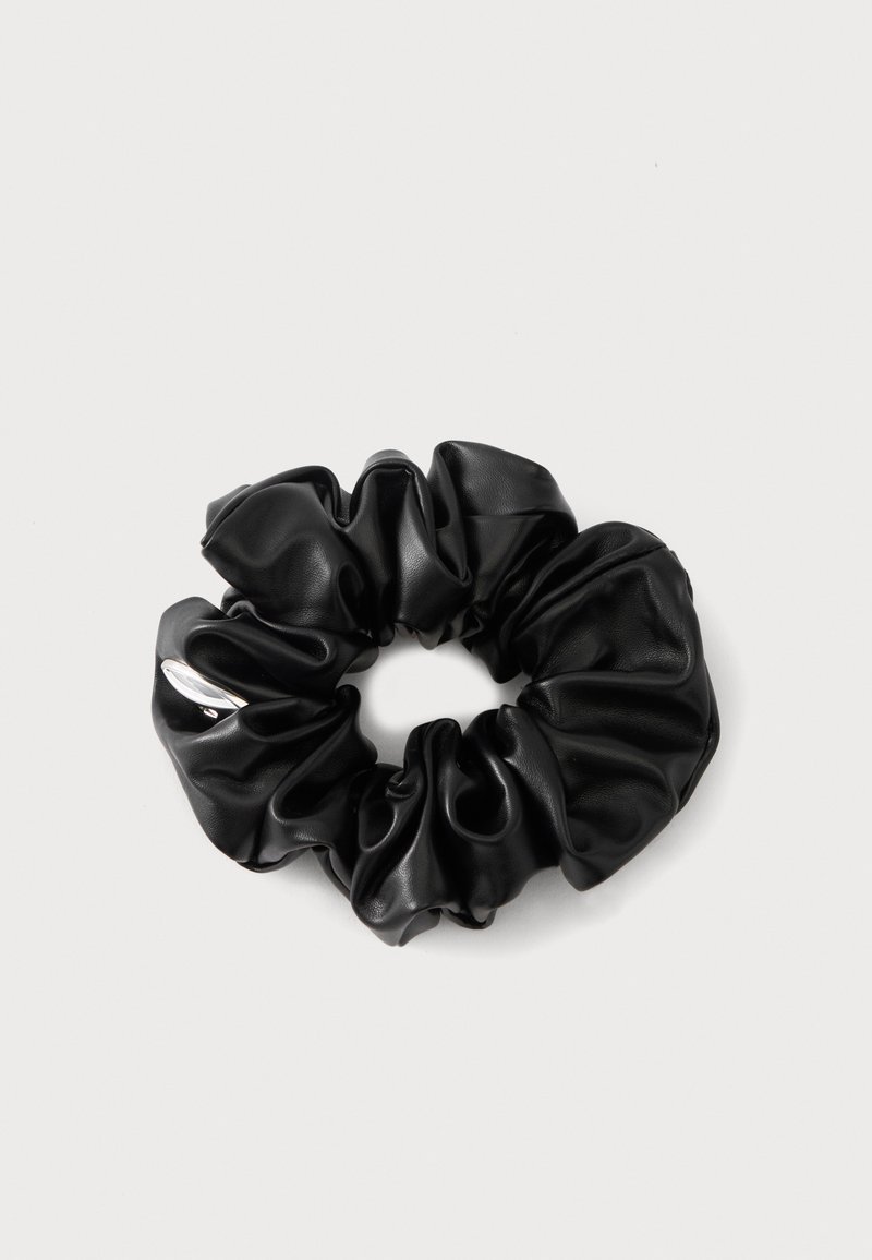 Chouchou noir brillant en tissu similicuir froncé avec une petite étiquette argentée sur fond gris clair.