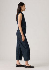 Dongerijumpsuit i mørkeblå med ærmeløs design, rett ben-kutt, frontlommer og minimale metalldetaljer. Kombinert med svarte flate sko.
