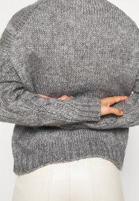 Šedý pletený svetr s ribovaným lemem a manžetami. Textura je silná s volným šitím, což ukazuje uvolněný design.