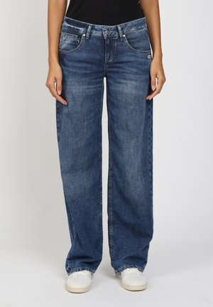 Front view of person wearing loose-fit blue jeans and white sneakers, standing against a plain white background.