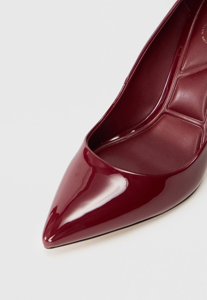Pump in vernice bordeaux con tacco alto, caratterizzata da un design a punta e una finitura liscia e lucida. L'interno presenta una fodera strutturata.
