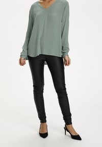 Blouse vert clair à manches longues avec un col en V et une texture subtile, associée à un pantalon noir moulant en similicuir et des escarpins noirs à bout pointu.