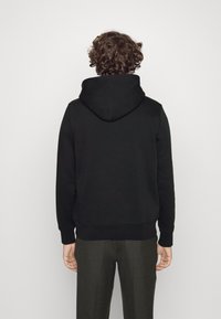 Sudadera con capucha negra hecha de una mezcla de algodón, que presenta un bolsillo tipo canguro y puños de canalé. La lleva con pantalones grises, mostrando un ajuste relajado.