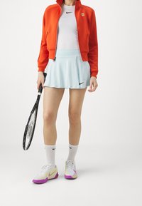 Röd jacka med dragkedja, vit Nike-tröja, ljusblå tennis kjol med svart logga, vita och lila sneakers, håller i en tennisracket.