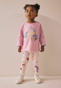 Een jong kind met krullend haar draagt een roze eenhoornshirt en een legging met patroon. Ze staat in een omgeving met neutrale kleuren en kijkt omhoog.