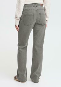 Persona vestida con jeans de pierna ancha grises y un suéter beige, de pie con la espalda hacia la cámara sobre un fondo blanco.