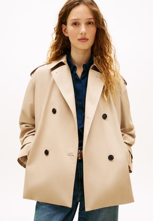 Jeune femme aux longs cheveux bouclés portant un trench-coat beige à double boutonnage sur une chemise bleu foncé et un jean bleu, les mains dans les poches.