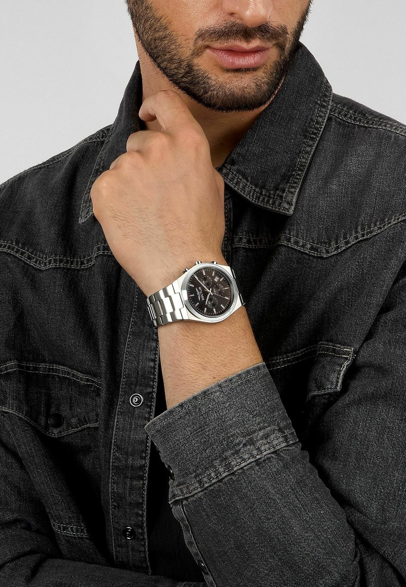 Orologio in acciaio inossidabile con cinturino tono argento, quadrante nero e dettagli tono argento. Indossato su una mano con una giacca di jeans scura.