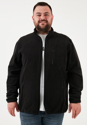 Chaqueta de forro polar negra con cremallera frontal, cuello alzado y bolsillos laterales. Presenta una textura suave y detalles de diseño mínimos.