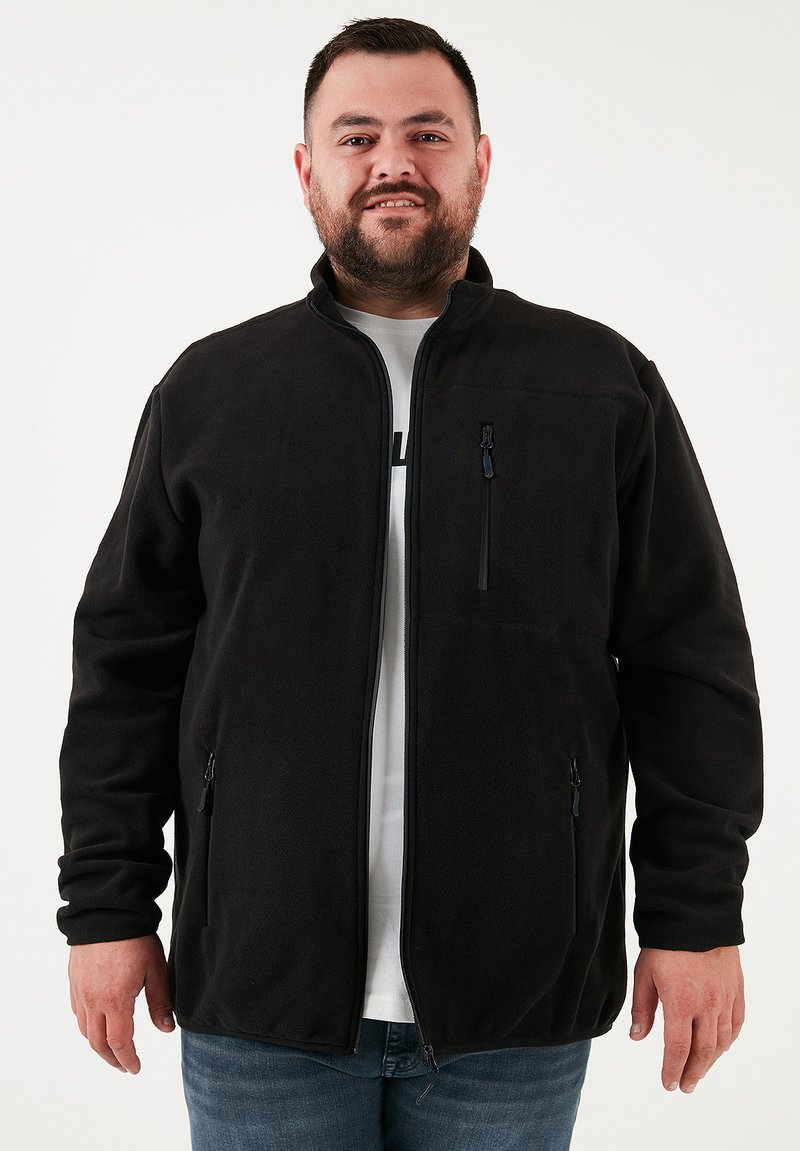 Chaqueta de forro polar negra con cremallera frontal, cuello alzado y bolsillos laterales. Presenta una textura suave y detalles de diseño mínimos.