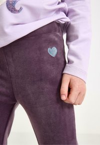 Leggings en velours violet avec une texture douce présentent un détail en broderie de cœur bleu clair sur la cuisse gauche. Associez-les avec un haut lavande.
