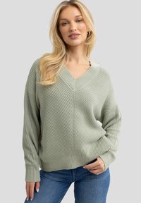 Lichtgroene V-hals trui met geribbelde textuur, lange mouwen en een relaxte pasvorm, gecombineerd met blauwe jeans voor een casual look.