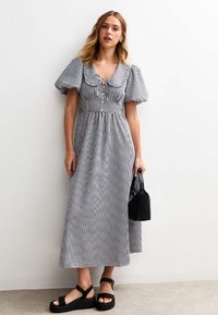 Robe maxi en vichy noir et blanc avec des manches bouffantes et un détail à nouer au niveau du col, associée à un petit sac à main noir et des sandales épaisses.