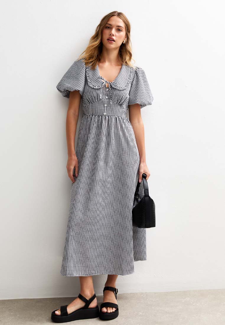 Robe maxi en vichy noir et blanc avec des manches bouffantes et un détail à nouer au niveau du col, associée à un petit sac à main noir et des sandales épaisses.