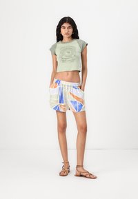 Top corto verde con design grafico abbinato a pantaloni shorts multicolori con motivi e accenti blu, arancione e verde. Sandali marroni.