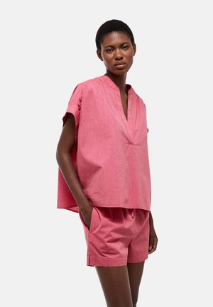 Femme portant un haut rose ample à manches courtes en V et un short assorti, debout avec une main dans la poche sur un fond blanc.