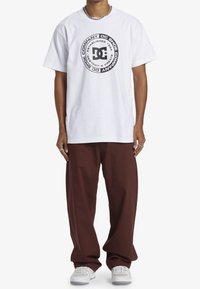 Weißes Baumwoll-T-Shirt mit einem schwarzen runden Logo, das den Text "DC Shoe Company" und das Gründungsjahr zeigt. Kombiniert mit lockeren braunen Hosen.