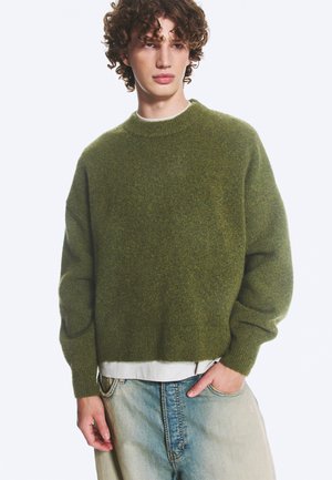 Jeune personne aux cheveux bouclés portant un pull vert duveteux sur une chemise blanche et un jean en denim bleu clair, debout avec une main dans la poche.