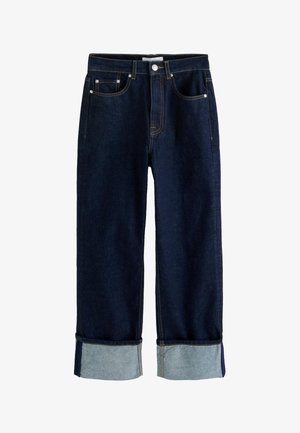 Kvinde med langt mørkt hår iført en lyseblå knap-skjorte, mørke jeans, store øreringe og holder en sammenfoldet mørk jakke mod en ensfarvet væg.