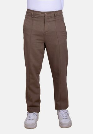 Hombre vestido con pantalones marrones de pierna recta y zapatillas blancas, de pie frente a un fondo liso.