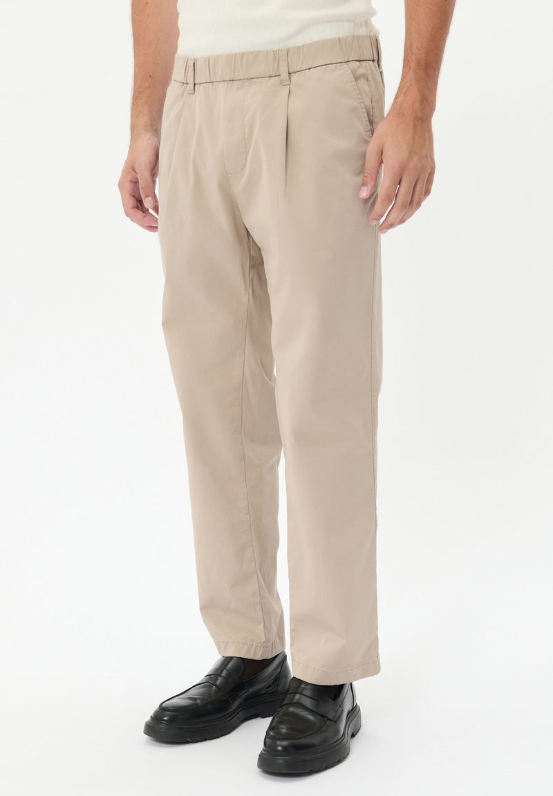 Pantaloni beige plissettati con vita elasticizzata e taglio dritto, abbinati a mocassini neri, su uno sfondo neutro.