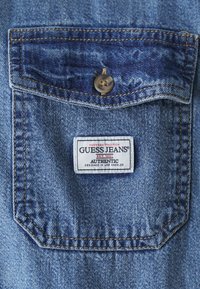 Denimhemd-Tasche mit Knopfverschluss, hellblauem Stoff mit verblasster Textur und einem Markenetikett mit rot-schwarzer Schrift.