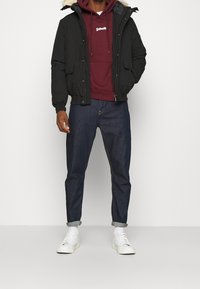 Czarna kurtka bomberka z kapturem wyściełanym futrem, bordowa bluza z kapturem z logo, ciemne jeansy denimowe i białe trampki. Gładka faktura, styl casual.