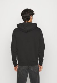 Uomo che indossa una felpa nera con cappuccio e jeans grigi scuri, in piedi rivolto verso un muro bianco semplice con la schiena rivolta verso l'osservatore.