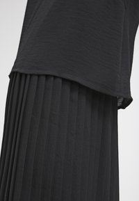 Jupe plissée noire avec un haut noir texturé superposé. Les tissus sont doux, avec une coupe décontractée et relaxed et sans fermetures visibles.