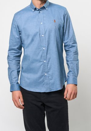 Hombre vistiendo una camisa azul claro ajustada con botones y un pequeño logo bordado en el pecho, acompañado de pantalones negros, sobre un fondo liso.