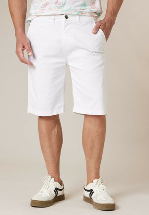 Homme portant un short blanc jusqu'aux genoux, une chemise à motifs floraux et des baskets blanches avec des accents noirs, debout les mains dans les poches.