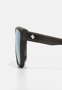 Juodų akinių nuo saulės storu rėmeliu, mėlynais lęšiais ir metaliniu logotipu ant šonkaulio. Dizainas pasižymi kampuotais kraštais ir blizgiu paviršiumi.