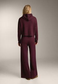 Hanorac tricotat scurt de culoare burgundă, cu glugă, asociat cu pantaloni evazați din același material și culoare. Textură fină și croială lejeră.