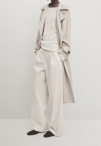 Trench-coat beige clair avec grand col, associé à un pull crème et un pantalon large crème. Des chaussures pointues foncées complètent la tenue.