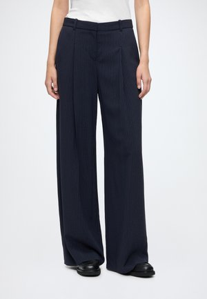 Person wearing navy blue wide-leg pinstriped trousers with black shoes and a white top, standing against a plain background.