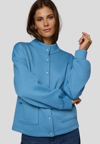 Blue quilted jacket with a high collar, button front, and side pockets. Features textured fabric and dropped shoulders for a relaxed fit.