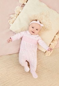 Lyserød baby romper med lange ærmer og blomster mønster. Hvid krave med lyserøde accenter. Blød tekstureret pude med tovværk i baggrunden.
