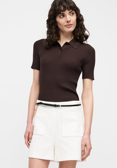 Femme aux cheveux bruns bouclés portant un polo marron foncé cintré à manches courtes et un short blanc taille haute avec de grandes poches et une ceinture noire.