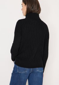 Pull en col roulé noir avec texture côtelée et motifs en losange. Associé à un jean en denim bleu. Le tissu semble doux et confortable.
