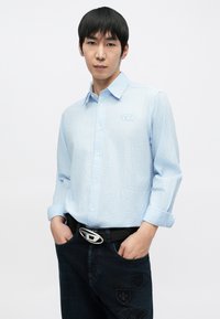 Világoskék, gombos ing finom csíkmintával és a mellkason hímzett logóval, sötét farmerral és ezüst övcsattal párosítva.