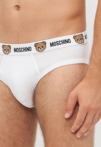 Witte katoenen slip met een zwart en bruin berenafbeelding op de elastische tailleband, gemarkeerd met "MOSCHINO" in vette letters.
