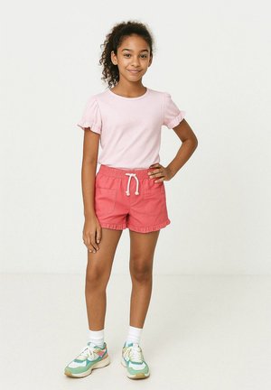Ung tjej med lockigt hår i en rosa blus med volanger, korallfärgade shorts med dragsko, vita strumpor och färgglada sneakers som står med handen på höften.
