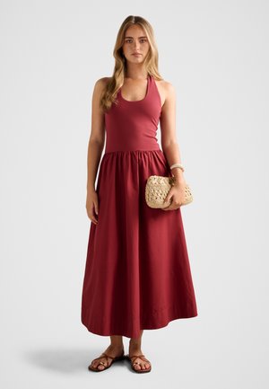 Fiatal nő hosszú szőke hajjal, piros ujjatlan nyakpántos ruhában, kezében fonott bézs táskával, és barna szandált visel.