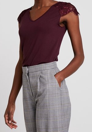 Frau trägt ein ärmelloses, dunkelviolettes Oberteil mit Spitzenärmeln und hoch Taillierte graue karierte Hosen, Hand in der Tasche.