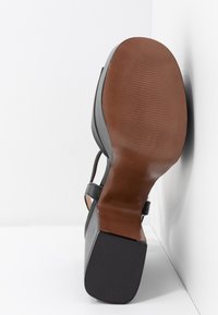 Sandale noire à talons hauts avec une fine bride autour de la cheville, dotée d'une semelle en cuir marron et d'une semelle extérieure en caoutchouc noir texturé pour une meilleure adhérence.