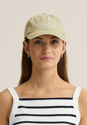 Ung kvinde med glat brunt hår iført sølv øreringe i hoop-design, en beige kasket med mærket "NEWHOUSE" og en hvid tanktop med sorte vandrette striber.
