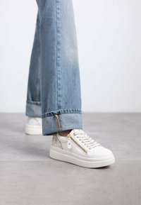 Zapatillas blancas texturizadas con un zipper lateral, herrajes dorados y un acento estampado. Combinadas con pantalones vaqueros cortados de color azul claro.