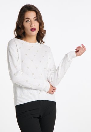 Jonge vrouw met golvend bruin haar die een witte trui met pareldetails en een zwarte broek draagt, poseren met één arm iets opgetild.
