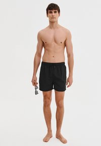 Czarne kąpielówki z elastycznym pasem i sznurkiem. Model trzyma czarne okulary przeciwsłoneczne, stoi boso na neutralnym tle.