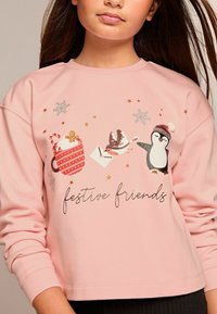 Sudadera rosa clara con motivos festivos: una taza roja, un bastón de caramelo, un pájaro y un pingüino, con acentos brillantes y el texto "amigos festivos".
